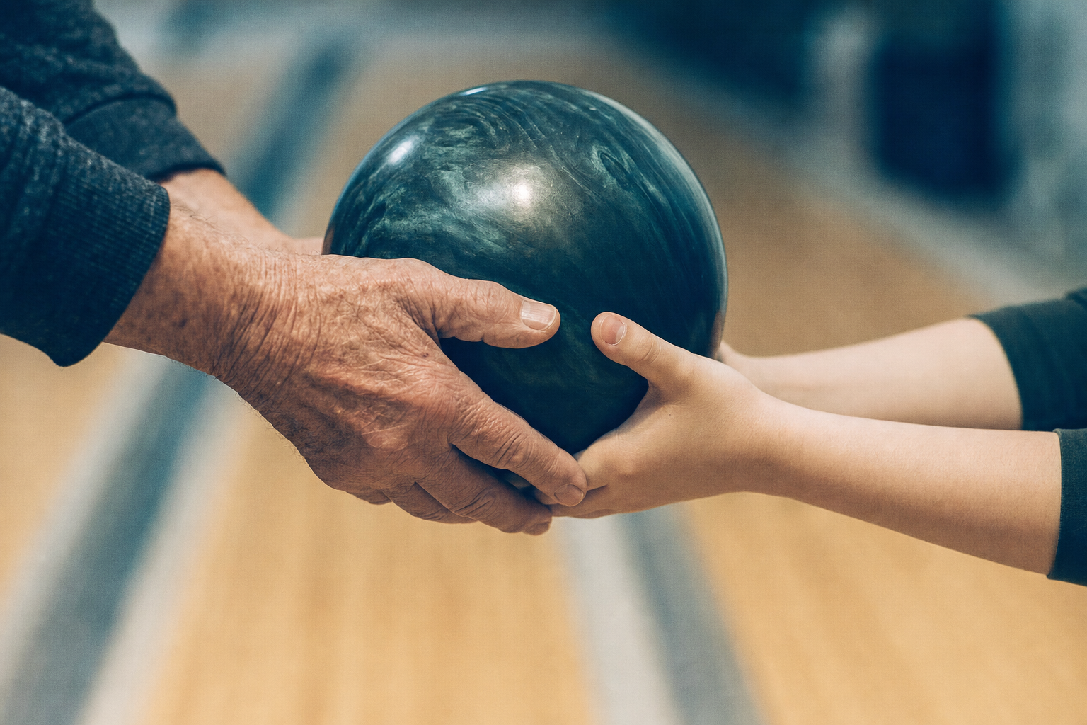 An older hand and a younger hand together holding a marbled bowling ball on a wooden lane.