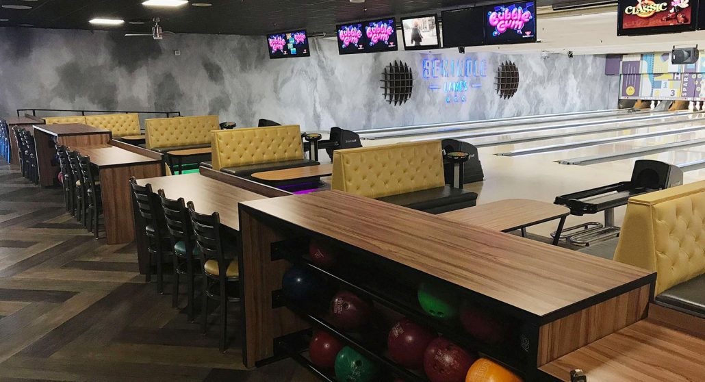 Interior of Seminole Lanes bowling center with wooden seating, bowling balls in racks, and the SEMINOLE LANES sign on the wall.